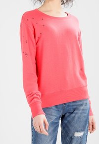 Felpa rosa corallo con maniche lunghe, scollo rotondo e piccoli dettagli grafici neri sulla spalla. Tessuto morbido, vestibilità rilassata.