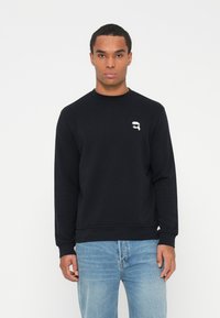 Svart crewneck genser laget av bomull, med en liten hvit logo på brystet og ribbestrikkede mansjetter og nederkant. Kommer sammen med lyseblå jeans.