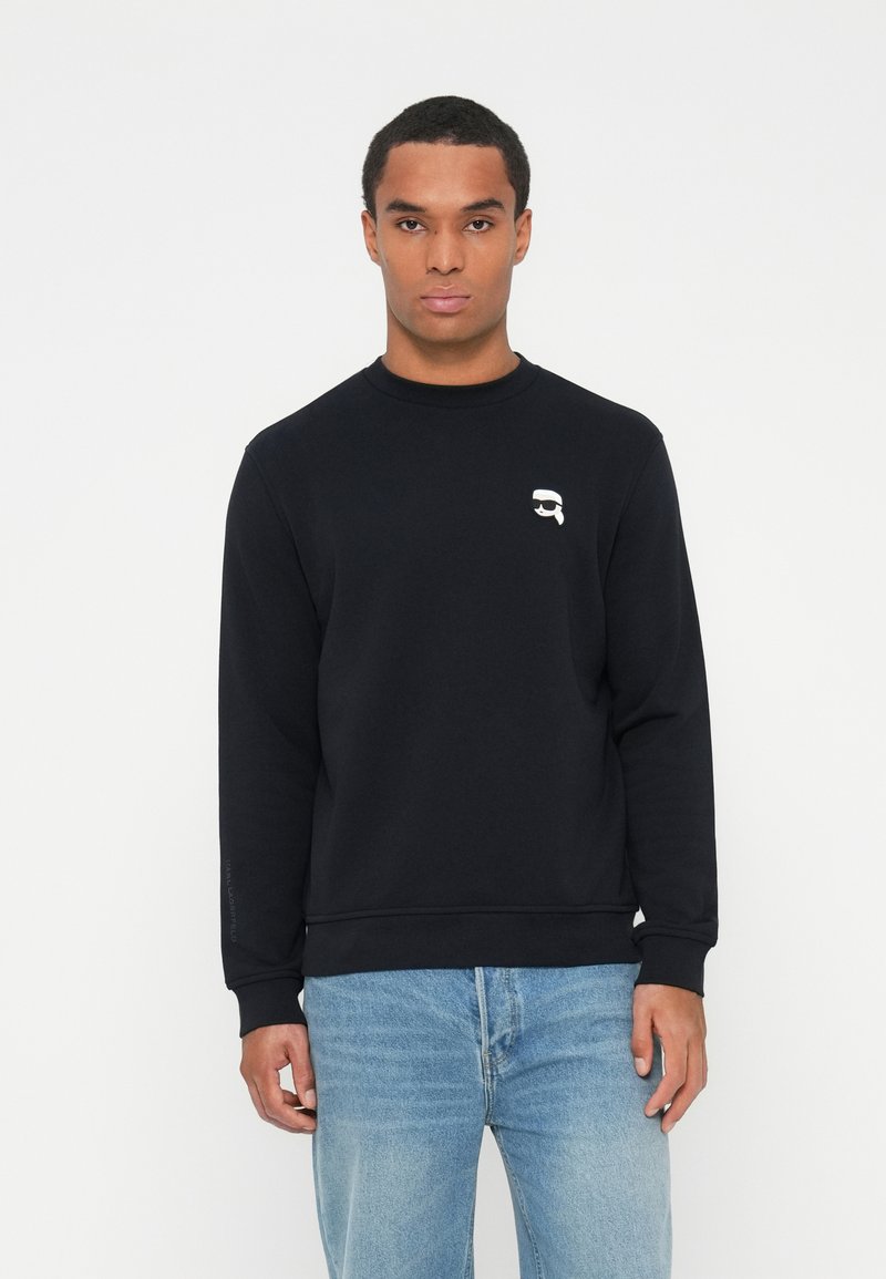 Svart crewneck genser laget av bomull, med en liten hvit logo på brystet og ribbestrikkede mansjetter og nederkant. Kommer sammen med lyseblå jeans.