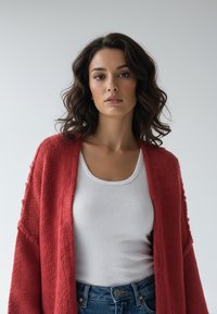 Czerwony dziergany kardigan na białym prążkowanym topie bez rękawów, charakteryzujący się luźnym krojem i teksturowaną tkaniną. Połączony z niebieskimi dżinsami.