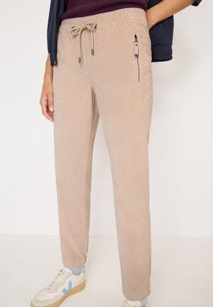 Beige, lichte broek met een elastische tailleband, verstelbaar trekkoord, zijzak met rits en taps tolopende benen. G gedragen met lichte sneakers.