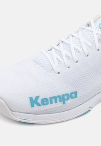 Hvit idrettssko med mesh-overdel og blå "Kempa"-logo, med avrundet tå, snøring og teksturert såle for grep.
