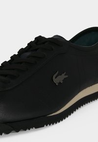 Μαύρο δερμάτινο sneaker με κορδόνια, ριγέ σόλα και μικρό μεταλλικό λογότυπο κροκοδείλου στο πλάι.
