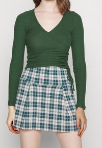 Grünes, geripptes langärmeliges Wickeltop, kombiniert mit einem weißen und teal-karierten, plissierten Rock. Der Rock hat eine Bindeschleife an der Taille.