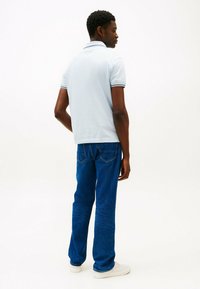 Camisa polo azul clara com colarinho e detalhe escuro, combinada com jeans retos azul escuro e ténis brancos, vista pela traseira.