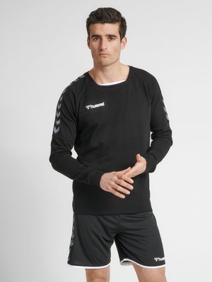 Sort sweatshirt med lange ærmer og rund hals, prydet med hvidt logo og chevron-detaljer på skuldrene; sat sammen med sorte shorts.