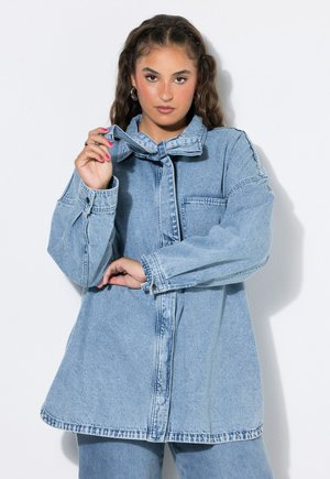 Kvinde med fletninger har en oversized lyseblå denimjakke og jeans på, holder en af jakke-kraverne op med den ene hånd.