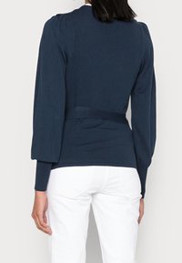 Maglione a maglia blu navy con maniche lunghe a palloncino, vita aderente e dettaglio con fiocco sulla schiena. Indossato con jeans bianchi, texture liscia.