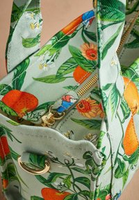 Kvetinová kabelka s zelenou tkaninou, na ktorej sú vzory v oranžovej, žltej a modrej farbe. Obsahuje zlatý zips a kovový detail s okom.