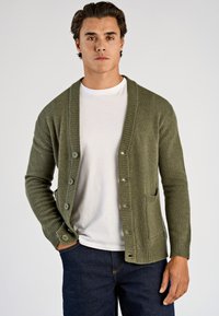 Olijfgroene cardigan met een geribde kraag, knoopsluiting, borstzakken en lange mouwen. Draagt over een wit T-shirt en donkere spijkerbroek.