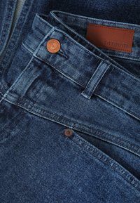 Nærbillede af foldede blå denimjeans med synlige bæltestropper, kobberknapper og brunt lædermærke med mærket "TOTUUM".