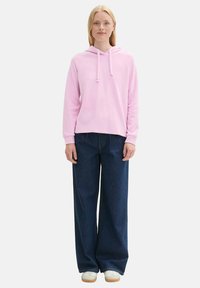 Svetloružová mikina s kapucňou a šnúrkami, kombinovaná s voľnými tmavomodrými džínsami a bielymi teniskami. Čistý, jednoduchý dizajn.