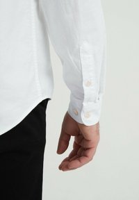 Chemise blanche à manches longues avec des poignets boutonnés, présentant une texture de tissu lisse et un ourlet arrondi. Main portant un pantalon noir présentée.