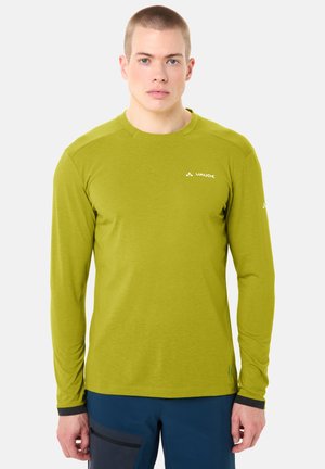 Uomo che indossa una maglia a maniche lunghe verde lime con il logo "VAUDE" e pantaloni blu scuro, in piedi contro uno sfondo bianco uniforme.