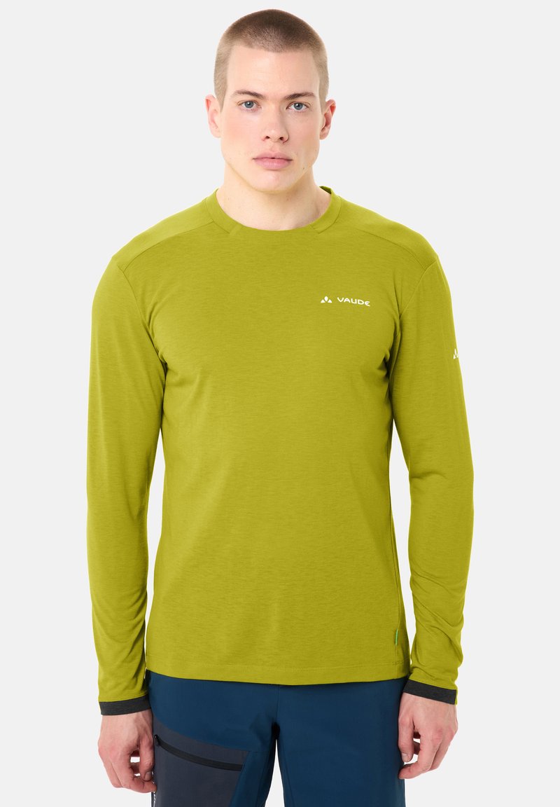 Homme portant une chemise à manches longues vert citron avec le logo "VAUDE" et un pantalon bleu foncé, debout devant un fond blanc uni.