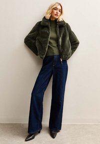Chaqueta de piel sintética verde con cuello, combinada con un suéter oliva y jeans de mezclilla oscuros de pierna ancha; modelo posando frente a un fondo neutro.