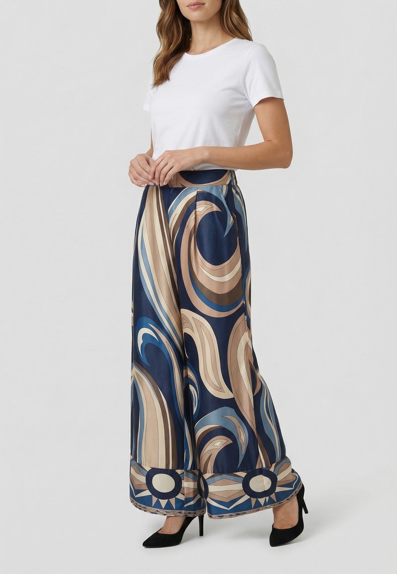 Donna che indossa una maglietta bianca a maniche corte e pantaloni a gamba larga con motivi swirl blu, beige e marroni, abbinati a tacchi alti neri.