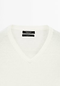 Pull en lin blanc à col en V avec étiquette "Massimo Dutti" à l'intérieur de l'encolure, fabriqué en Chine, présenté sur un fond uni.