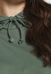 Sudadera con capucha verde con cuello fruncido y cordones ajustables. Tela suave con detalles texturizados en los extremos de los cordones.