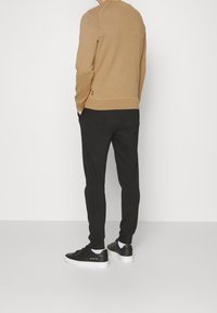 Beige gestrickter Sweatshirt mit gerippten Bündchen, kombiniert mit schwarzen Jogginghosen und schwarzen Turnschuhen mit weißer Sohle.