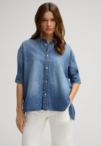 Light blue denim shirt with a button-down front, short rolled sleeves, and a curved hem. Textured fabric with subtle fading details.