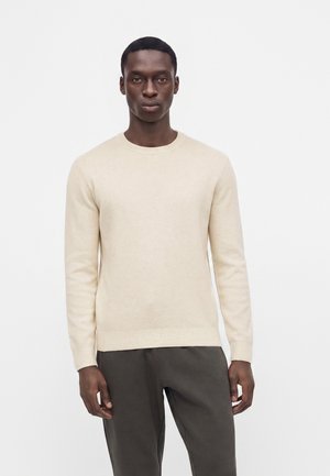 Homme debout sur un fond blanc, portant un pull beige à manches longues et un pantalon gris foncé, faisant face à l'avant avec une expression neutre.