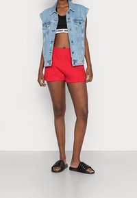 Shorts rouges avec une ceinture élastique, un crop top noir et une veste sans manches en denim. Des sandales à semelles nervurées noires complètent la tenue.