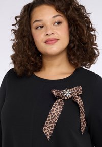 Czarna sukienka ozdobiona kokardą w panterkę z dodatkiem kamienia szlachetnego, umieszczoną na lewym ramieniu, wykonana z teksturowanej tkaniny i z okrągłym dekoltem.