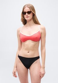 Γυναίκα με γυαλιά ηλίου τύπου cat-eye σε σχήμα οστρακόδερμου, τοπ μπικίνι κοραλί με λευκή περίμετρο και μαύρο κάτω μέρος μπικίνι μπροστά σε απλό φωτισμένο φόντο.