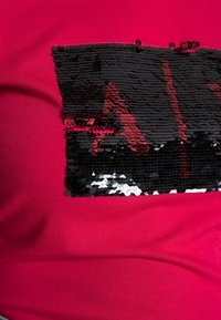 Top de algodón fucsia con un parche de lentejuelas negras y acentos rojos. Las lentejuelas crean una superficie texturada y reflectante.