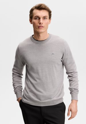 Giovane uomo che indossa un maglione girocollo grigio chiaro e pantaloni neri, in piedi con una mano in tasca contro uno sfondo neutro.