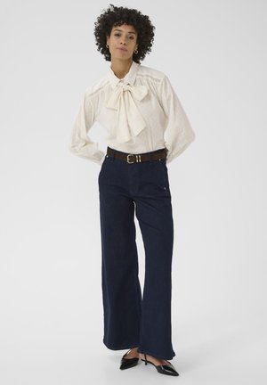 Weiße Bluse mit Schleife, strukturiertem Muster und lockeren Ärmeln. Marineblaue weit geschnittene Jeans. Brauer Gürtel mit goldener Schnalle und schwarzen spitzen Schuhen.