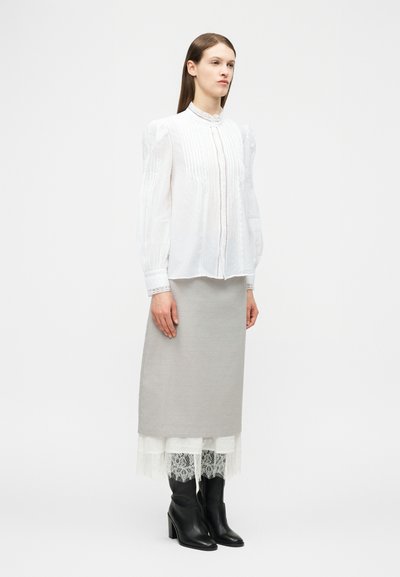 Γυναίκα που φοράει λευκό μπλουζάκι με ψηλό γιακά, ανοιχτό γκρι midi φούστα με δαντέλα και μαύρες μπότες με τακούνι, που στέκεται μπροστά σε ένα απλό φόντο.