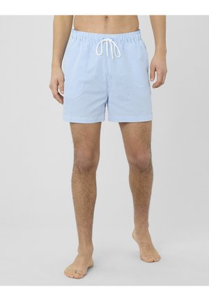 Homme portant un short de bain rayé bleu clair et blanc avec une taille élastique et un cordon de serrage blanc, debout pieds nus sur un sol gris.
