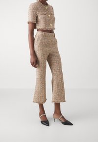 Kvinna klädd i en beige tweed kortärmad jacka med guldknappar och matchande högmidjade, korta utsvängda byxor, svarta klackskor med silverremmar.