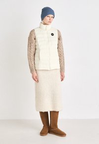 Crème puffer vest met een hoge kraag, gecombineerd met een beige gebreide trui, fluffy crèmekleurige rok en bruine suède kniehoge laarzen.