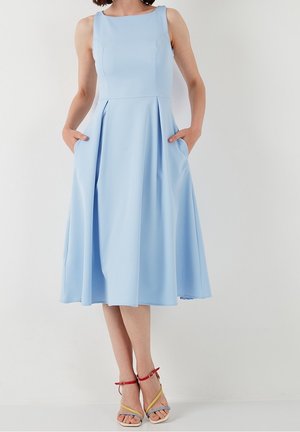 Woman wearing sleeveless light blue midi dress with pockets, paired with multicolored high-heeled sandals, standing against a white wall.