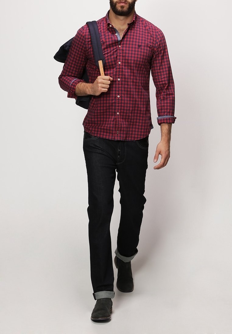 Homme portant une chemise à carreaux rouge et noir, un jean noir retroussé aux chevilles, des chaussures noires, une veste bleu marine sur l'épaule.