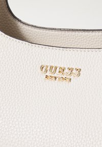 Sac à main en cuir texturé blanc avec le logo doré "GUESS" et l'inscription en relief "EST 1981". Il présente un bord supérieur lisse et incurvé ainsi que de subtils détails de couture.