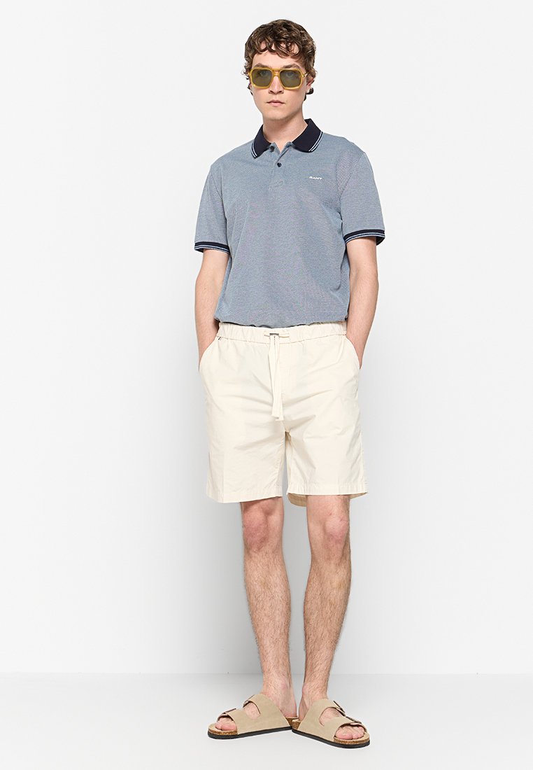 Poloshirt in lichtblauw met een marineblauwe kraag, witte shorts met koord, beige sandalen en gele zonnebril. Casual zomeroutfit.