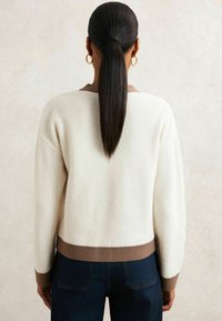 Sweter w kolorze kremowym, wykonany z fakturowanej tkaniny, z brązowymi ściągaczami przy mankietach i dolnej krawędzi. Stylizowany z ciemnoniebieskimi dżinsami, widok z tyłu.