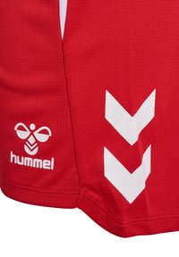 Shorts de sport rouges avec logo d'abeille Hummel blanc et deux bandes chevrons blanches descendantes sur le côté.