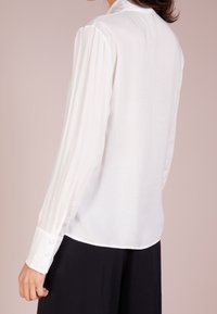 Blouse blanche à manches longues avec une finition texturée, coupe ajustée et poignets boutonnés, dotée d'un col classique et d'une drape fluide.
