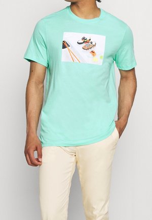 Mężczyzna noszący jasnorośli t-shirt z grafiką sushi i sneakersów, zestawiony z beżowymi spodniami na jednolitym tle.