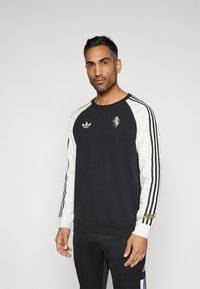 Črna jopa z belimi rokavi, na kateri je logotip Adidas in majhen vezen motiv. Kontrastni trakovi na rokavih, mehka tekstura.