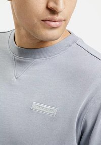 Jasnoszara bluza z ribbingu przy okrągłym dekolcie, z małą teksturowaną naszywką z napisem "CROSS JEANS" na piersi. Miękka struktura materiału.