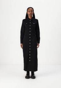 Robe en denim noir avec des manches longues, fermeture �à bouton sur le devant, deux poches poitrine et poches latérales. Le modèle porte des bottes noires chunky.