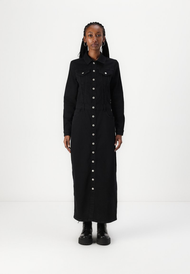 Robe en denim noir avec des manches longues, fermeture à bouton sur le devant, deux poches poitrine et poches latérales. Le modèle porte des bottes noires chunky.