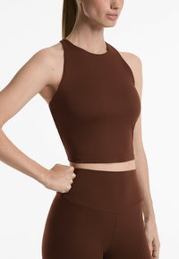 Crop top maro, ajustat, cu un gât înalt și design fără mâneci, realizat dintr-un material neted, asortat cu pantaloni înalți.