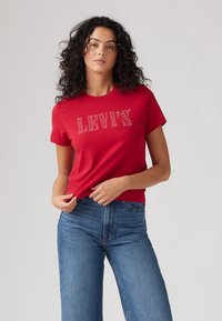 Punane puuvillane T-särk, millel on särava kiviga "LEVI'S" logo, koos kõrge vöökohaga helesiniste teksadega, näidates lõdva istuvust.